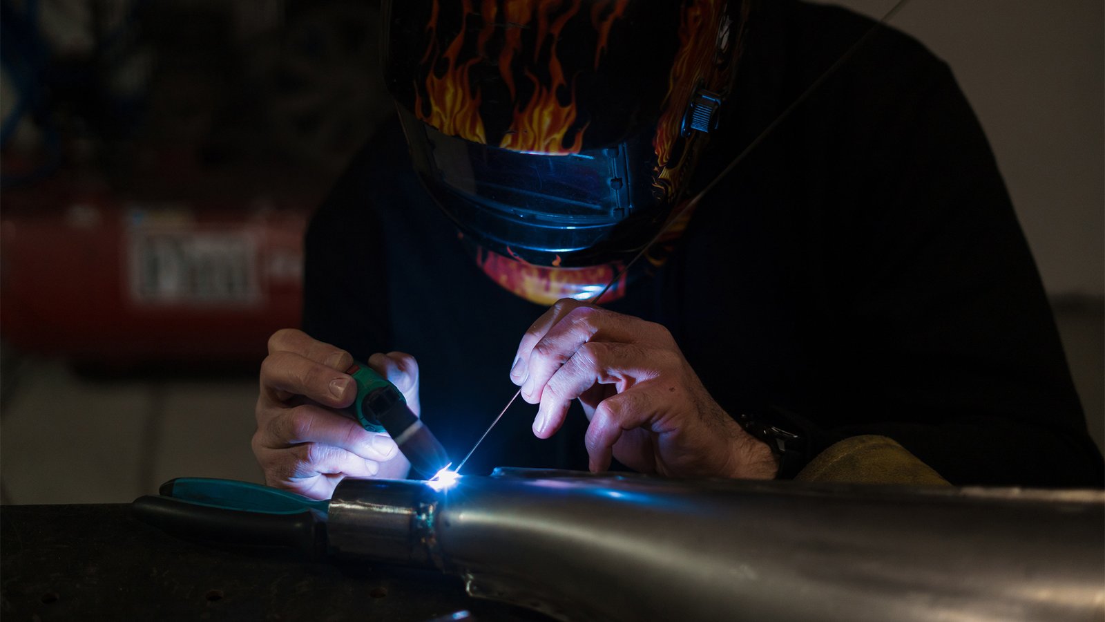 Worker in helmet with flames welding the steel part of a motorbike using an argon welding machine and gloves in a garage